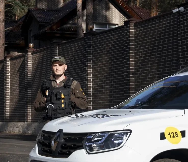 Охоронець біля високого цегляного паркану садиби, поруч службовий автомобіль
